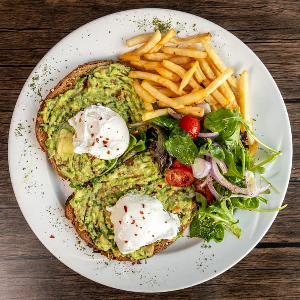 Avocado toast with poached eggs, a side of french fries, and a fresh salad on a white plate.