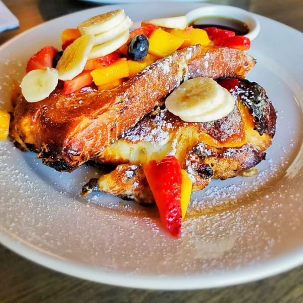 Golden French toast topped with powdered sugar, sliced bananas, strawberries, mango, and blueberries.